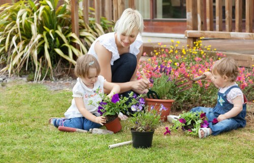 A woman with short blond hair and a white blouse is sitting on a well-maintained grassy lawn in a garden, engaging with two young children who are planting and tending to potted flowers. The children, both with light-colored hair, are wearing casual summer clothes: one in a white t-shirt and jeans, the other in a patterned dress. The garden features a lush lawn bordered by a variety of flowering plants and shrubs, with a wooden deck or patio area visible in the background. There is a mix of colorful blooms, including purple, pink, and yellow flowers, enhancing the vibrant, natural setting. A garden trowel is lying on the grass near the children, suggesting active gardening. The environment appears bright and sunny, with natural light illuminating the scene. This outdoor space exemplifies a well-kept, family-friendly garden, suitable for gardening, outdoor leisure, and children’s play, reflecting natural tones, plant textures, and garden maintenance typical of outdoor gardening services in Chigwell, Essex, highlighting the expertise of Hedge Trimming Chigwell in garden care.